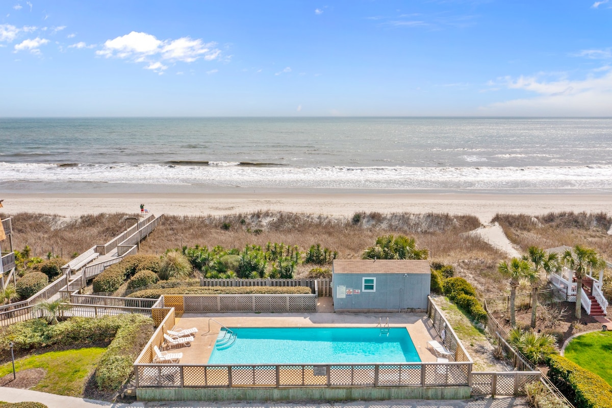 An expansive view captures a clear blue sky meeting the ocean's horizon. The beach features sandy areas and subdued vegetation. A pool is visible below, framed by a wooden deck and lounge chairs, providing a tranquil gathering spot near the waterfront.