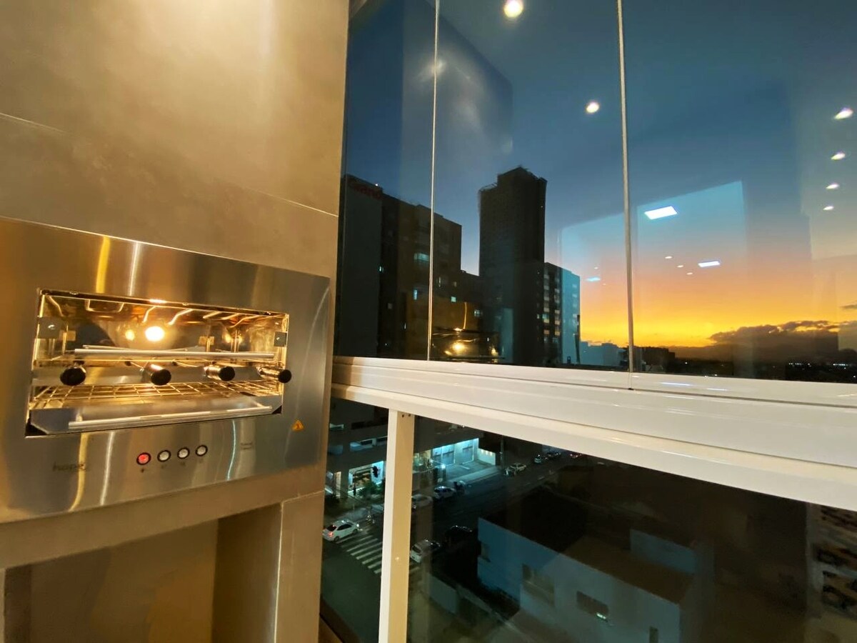 An electric grill is mounted on a kitchen wall, featuring controls and a stainless steel finish. A large window offers a view of the city skyline at sunset, with soft hues of orange and pink illuminating the sky.