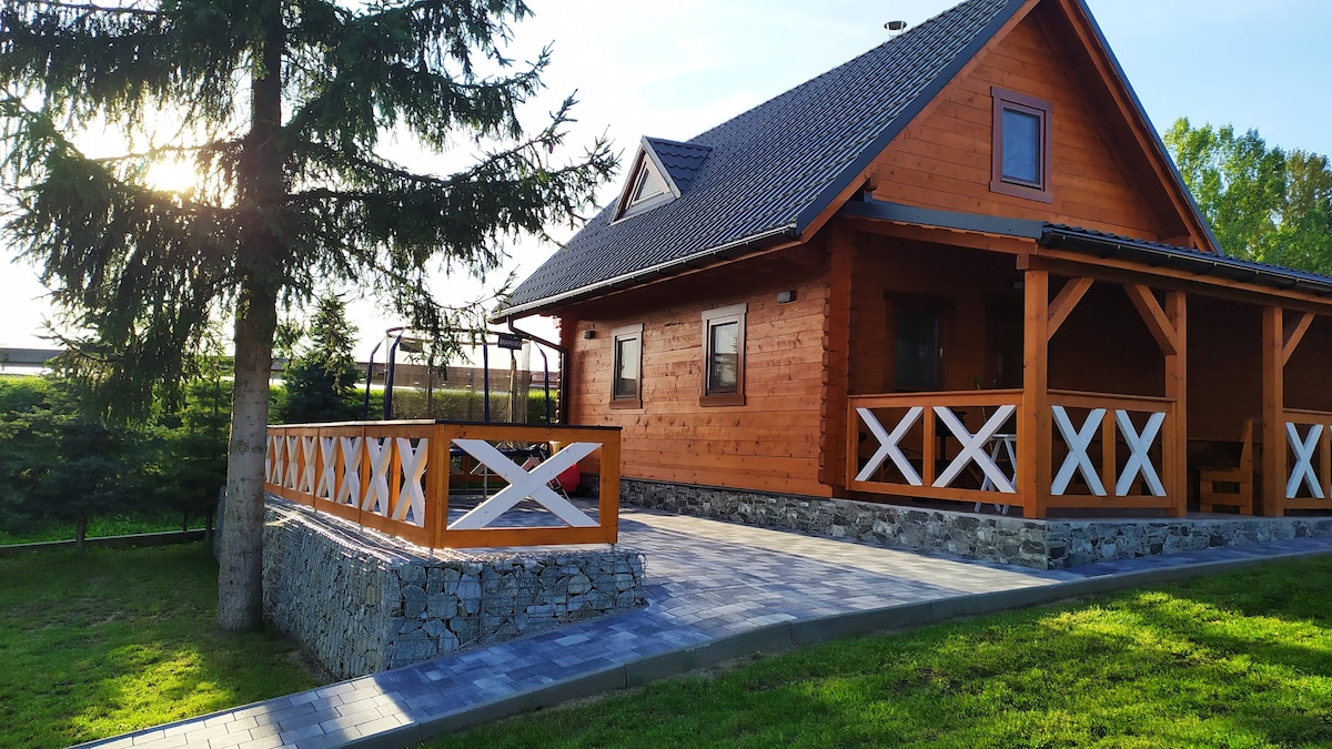 A charming wooden house is set against a sunny backdrop, featuring a spacious patio with a stone wall. The wraparound porch is supported by wooden beams, and a green lawn with a tall tree adds to the serene setting.