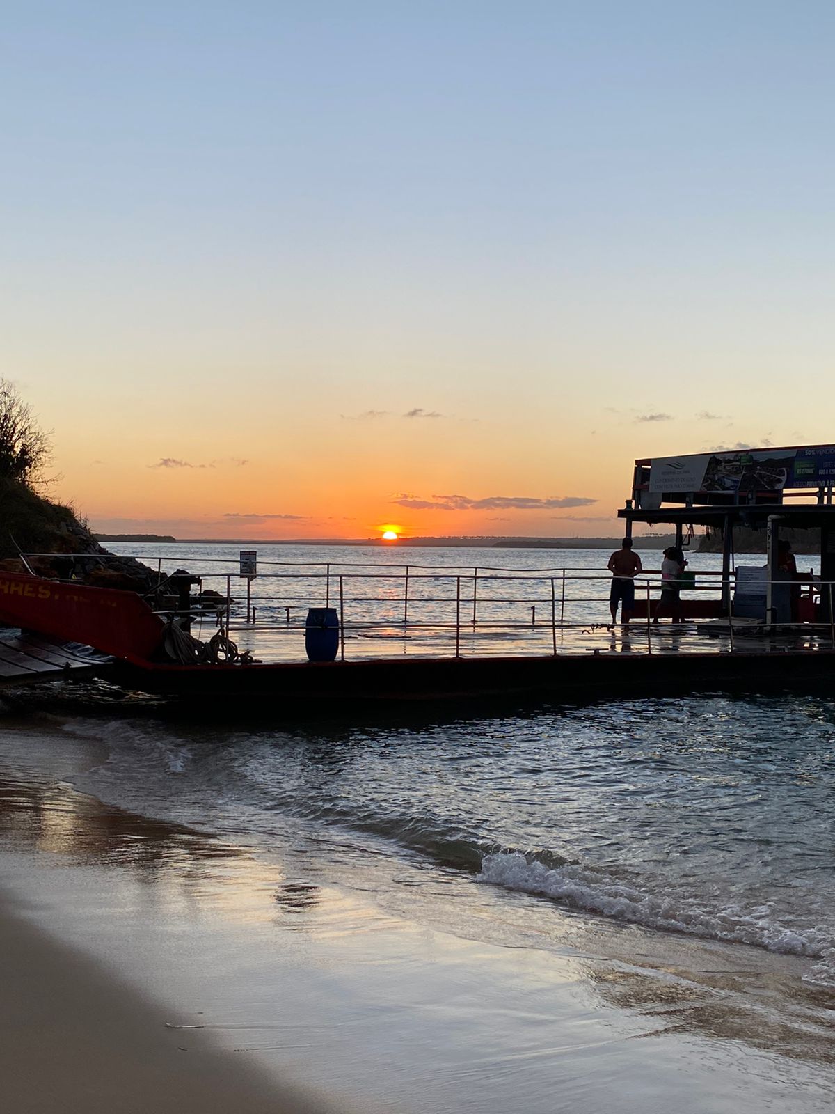 The serene beach scene captures a sunset over the water, with warm hues painting the sky. A calm shoreline gently meets the waves. A structure is visible at the water's edge, with a person standing nearby and silhouetted against the vibrant backdrop.