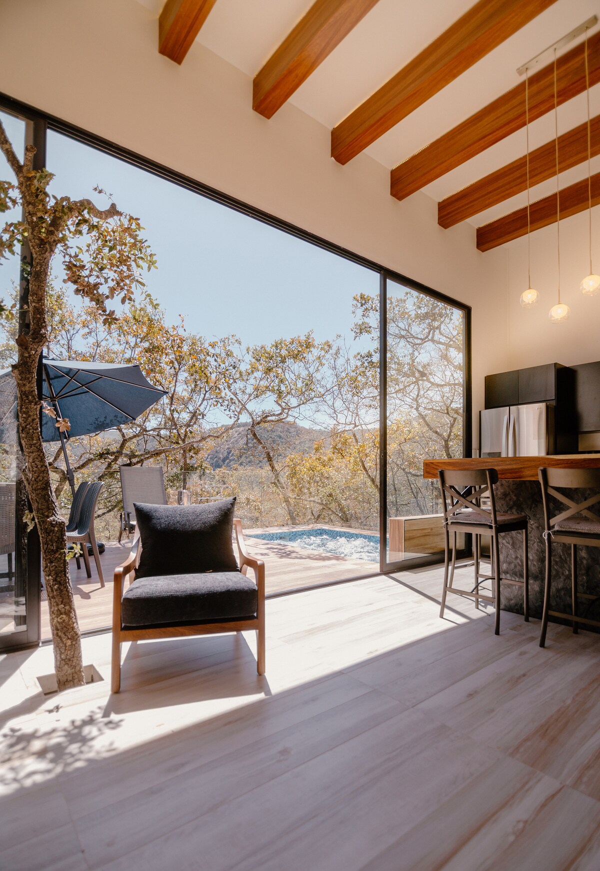 Large windows provide a view of the outdoor area, revealing a sunny terrace with a dining space, an umbrella, and a glimpse of a pool. Inside, a wooden dining bar sits beside a comfortable chair, with beams accentuating the ceiling design.