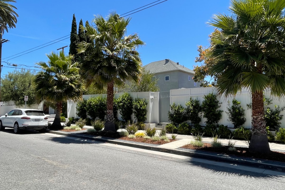 A street view showcases a neatly landscaped property surrounded by tall palm trees and lush greenery. A white fence encloses the area, adding to the sense of privacy. A light-colored vehicle is parked along the roadside, under a clear blue sky.