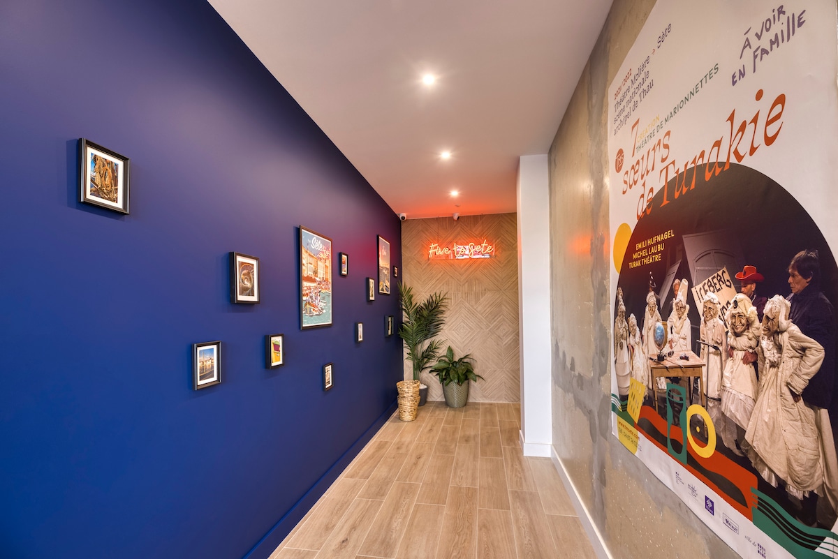 A hallway is presented with a deep blue wall featuring an arrangement of framed artwork. Soft lighting highlights the pieces, while a colorful mural on one side adds visual interest. A decorative plant is placed in a corner, contributing to the inviting atmosphere.