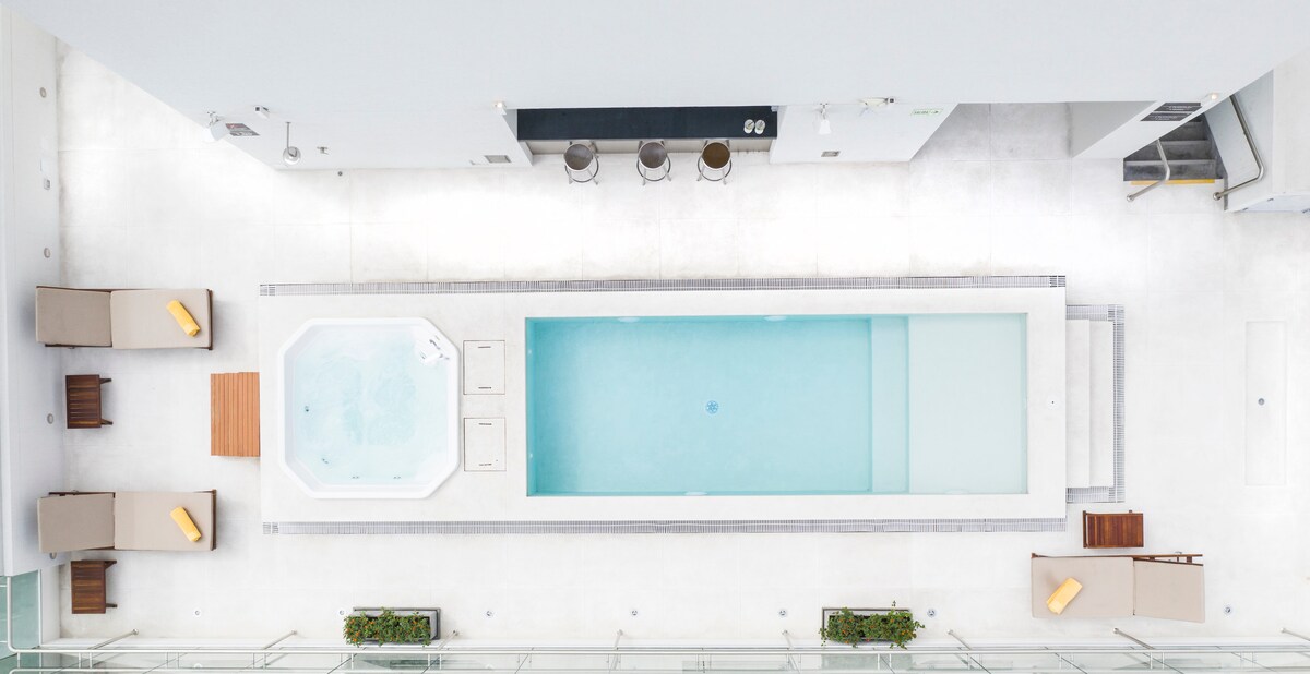 An aerial view of a rooftop area reveals a long, narrow pool and a hot tub, both surrounded by light-colored tiles. Loungers with soft cushions are positioned nearby, creating a relaxing atmosphere. A bar with high stools is visible along one side.