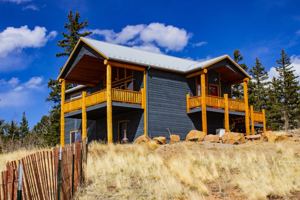 A modern cabin is positioned on a hillside, featuring large covered decks at the front. Natural wooden accents contrast with dark exterior walls. The landscape is dotted with grasses and rocks, while a clear blue sky with fluffy clouds is visible above.