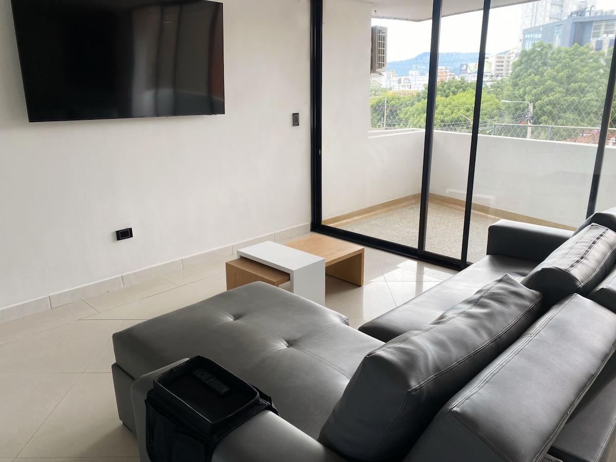 A spacious living area is displayed, featuring a comfortable grey sofa and a modern coffee table. A wall-mounted TV is visible, alongside large glass doors that provide a view of the city and natural light. The light-colored tiled floor enhances the open atmosphere.