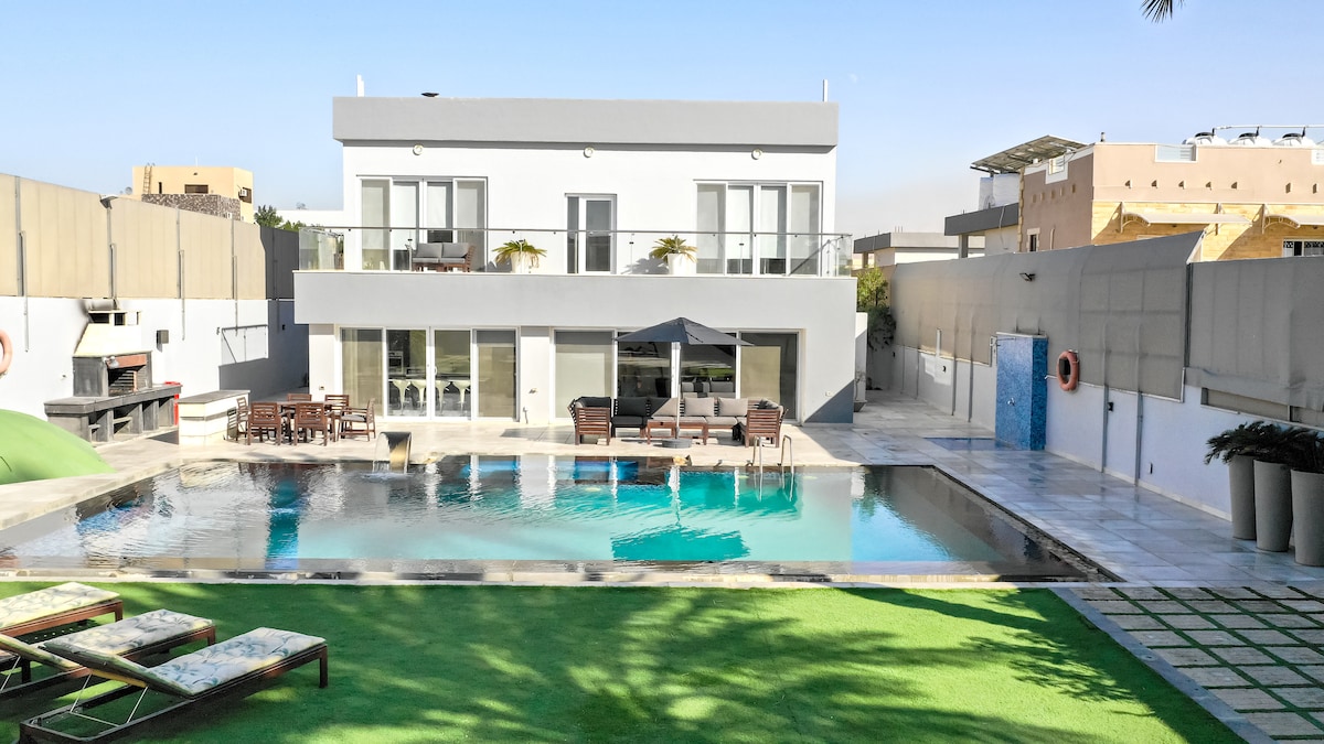 A modern villa is showcased with a spacious, inviting overflow pool reflecting the clear sky. Sun loungers are positioned on a manicured lawn, and outdoor seating is arranged by the poolside. Large glass doors provide an unobstructed view of the stylish interior.