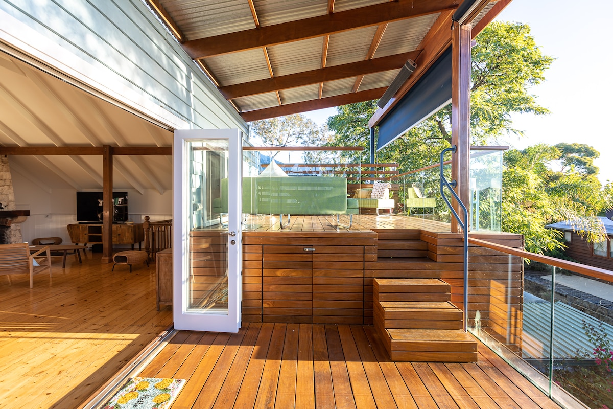 The outdoor deck features natural wood finishes and steps leading to a lower area. Glass railings provide unobstructed views of the surrounding trees. A folding door opens to seamlessly connect the indoor and outdoor spaces, promoting a flow of natural light.