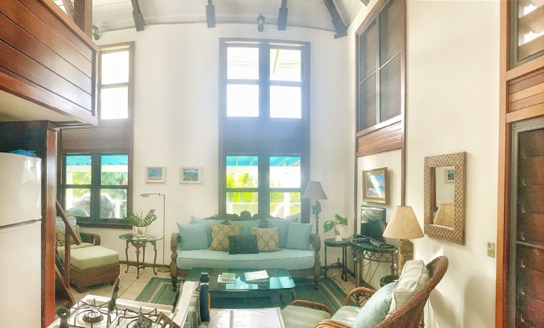 A light-filled living space features high ceilings and large windows, offering views of the surrounding lush greenery. A comfortable seating area contains a soft blue couch with decorative pillows and a coffee table. Warm wooden accents and various artworks enhance the inviting atmosphere.