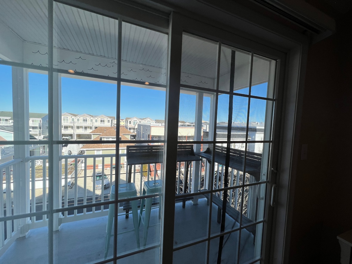 A covered balcony is visible, featuring a small table and two chairs, creating an outdoor seating area. Large sliding glass doors provide access and showcase a view of nearby buildings and a clear blue sky, allowing natural light to fill the space.