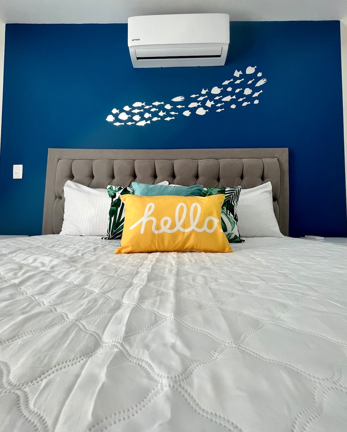 A spacious bed is centered against a deep blue wall, featuring a decorative fish motif above. The bed is dressed with crisp white linens and a bright yellow 'hello' pillow, complemented by green accents on either side.