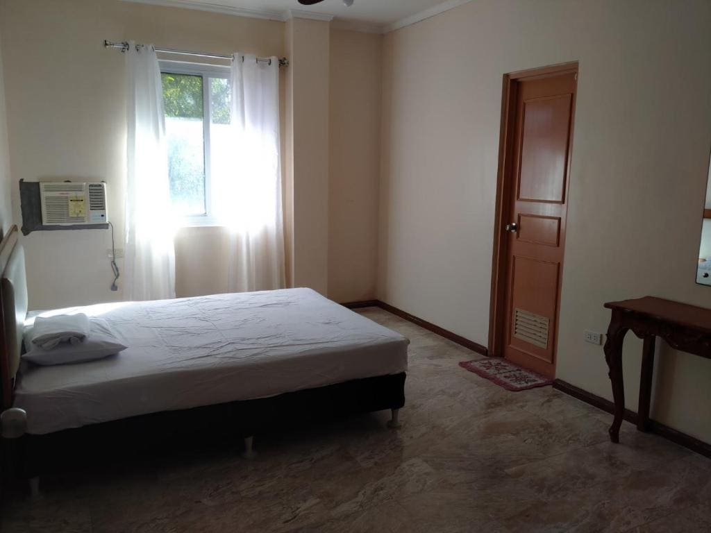 A well-lit bedroom features a spacious bed with a simple white bedspread. Natural light filters through a window adorned with sheer curtains. A small dressing table is positioned against one wall, while an air conditioning unit is mounted above the bed.