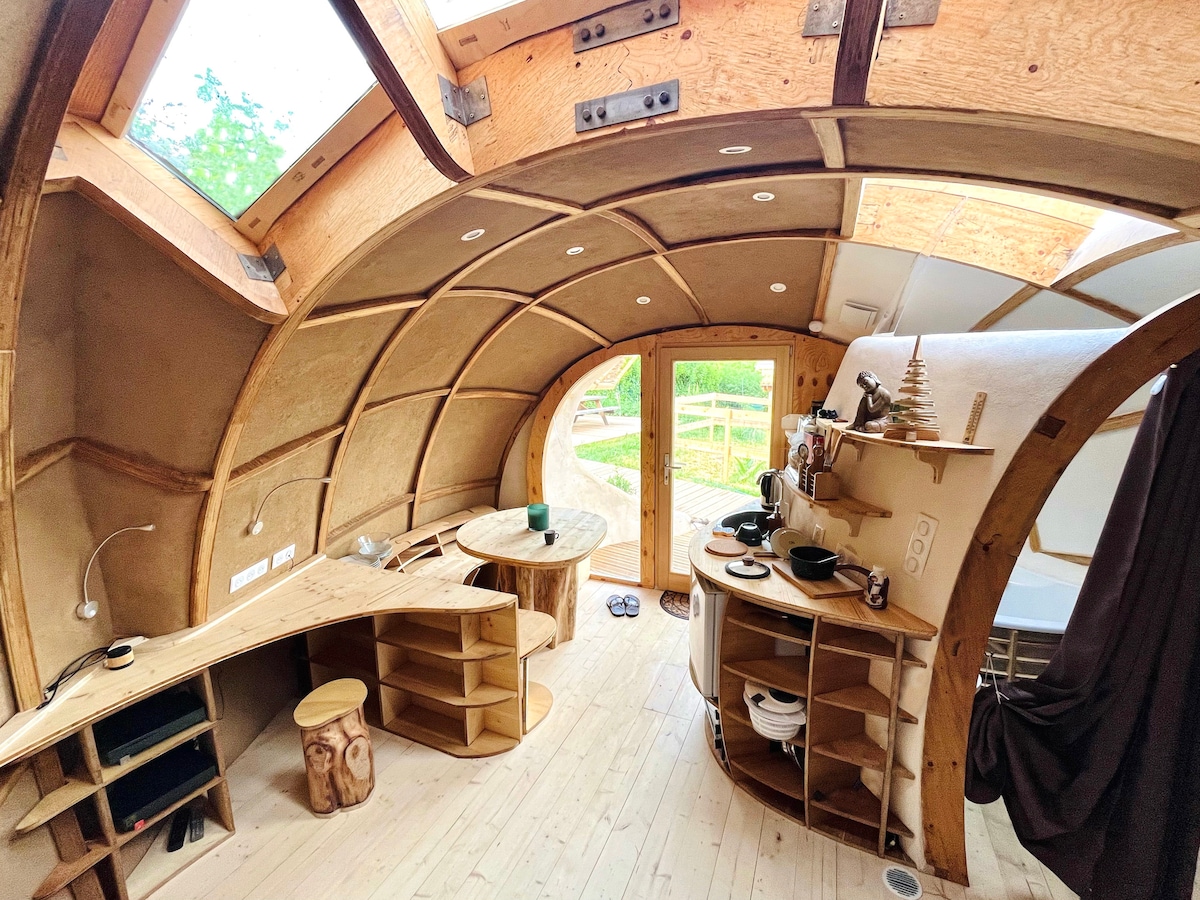 An interior view showcasing a semi-circular, wooden structure with earthy tones. Curved walls integrate shelving and hidden lighting. A small kitchen area features wooden shelving and a round table. Large windows line the ceiling, providing natural light and a view of the outdoors.