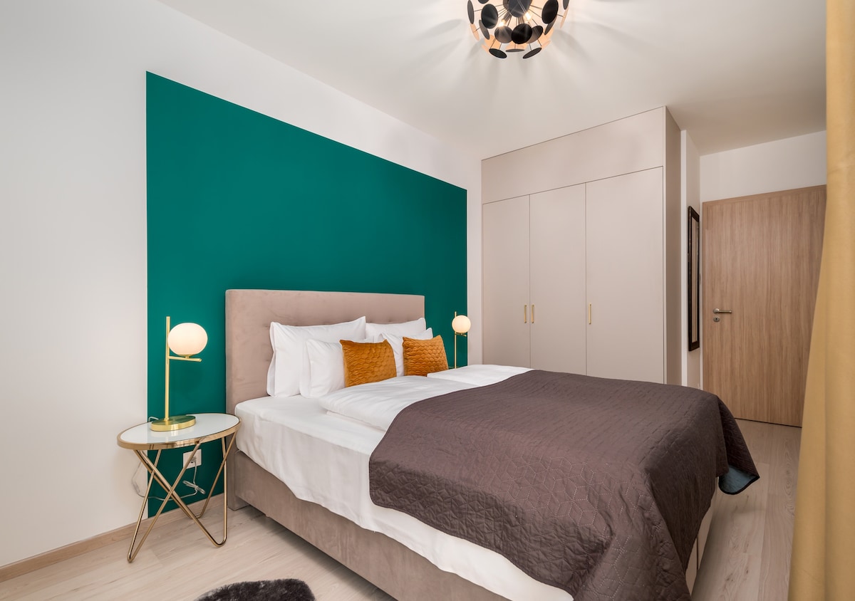 A modern bedroom features a spacious bed with white linens and decorative pillows against a deep green accent wall. A bedside table with a soft-glow lamp is positioned beside the bed. A stylish light fixture illuminates the room, enhancing the contemporary decor.