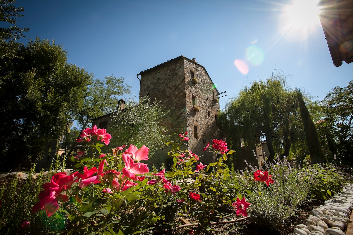 A historic stone building stands against a clear blue sky, surrounded by vibrant pink flowers in the foreground. Lush greenery frames the structure, with sunlight casting a gentle glow on the scene, highlighting the natural beauty of the Tuscan landscape.