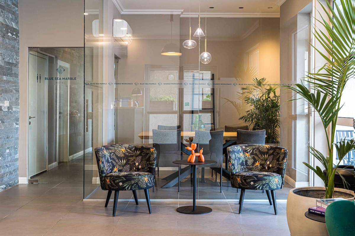 A modern lobby features a glass partition separating a dining area from a cozy seating space. Two patterned armchairs are positioned around a small black table, which displays decorative items. Natural light fills the room, with indoor plants adding a fresh touch.