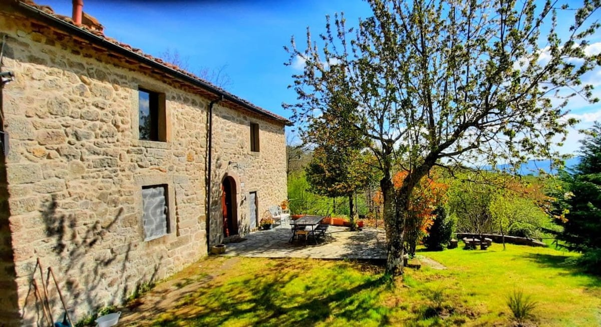 A rustic stone farmhouse is surrounded by lush greenery and well-maintained grounds. A spacious outdoor area features several wooden tables and chairs, perfect for dining or relaxation. The structure is complemented by trees and flowering plants, creating a serene natural setting.