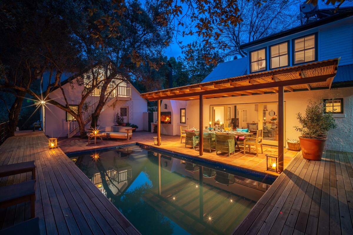 The outdoor area features a tranquil swimming pool surrounded by a wooden deck, illuminated by soft lighting. Nearby, a spacious dining table is set under a covered patio, with views of the house and surrounding trees enhancing the inviting ambiance.