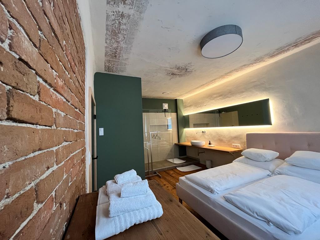 A modern bedroom features a light-colored, spacious bed with soft bedding. Exposed brick walls add texture, while a sleek bathroom area is visible with a glass shower and large sink. Natural wood elements, such as a simple table with neatly arranged towels, enhance the design.