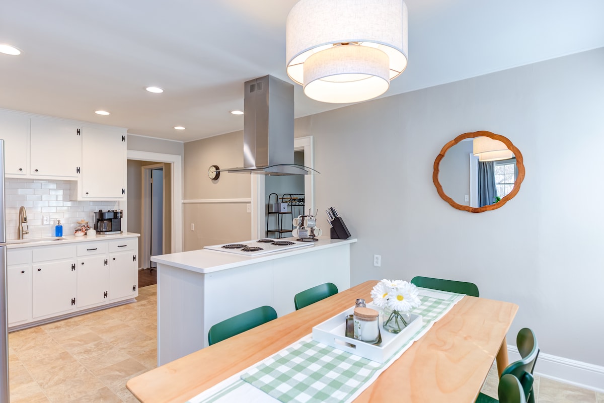 A modern kitchen and dining space features a light wooden table set with a green and white checkered runner. The area is illuminated by a stylish pendant light, with sleek cabinets and a contemporary range visible in the background.