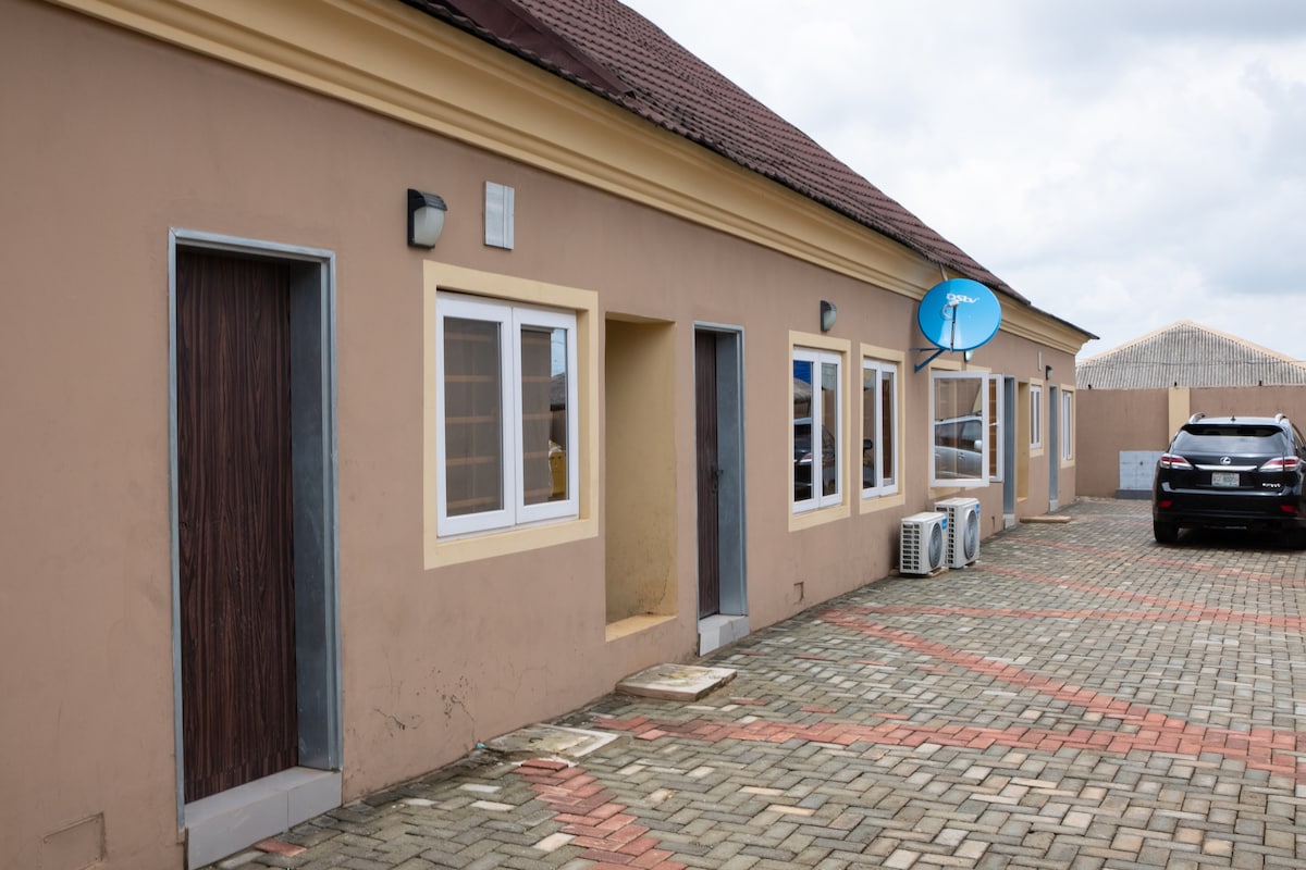 The exterior of the building is showcased, featuring a smooth, tan facade with a decorative trim. Windows are aligned along the walls, allowing natural light. A parked vehicle and air conditioning units are visible on the stone-paved pathway, creating a practical and accessible entrance.