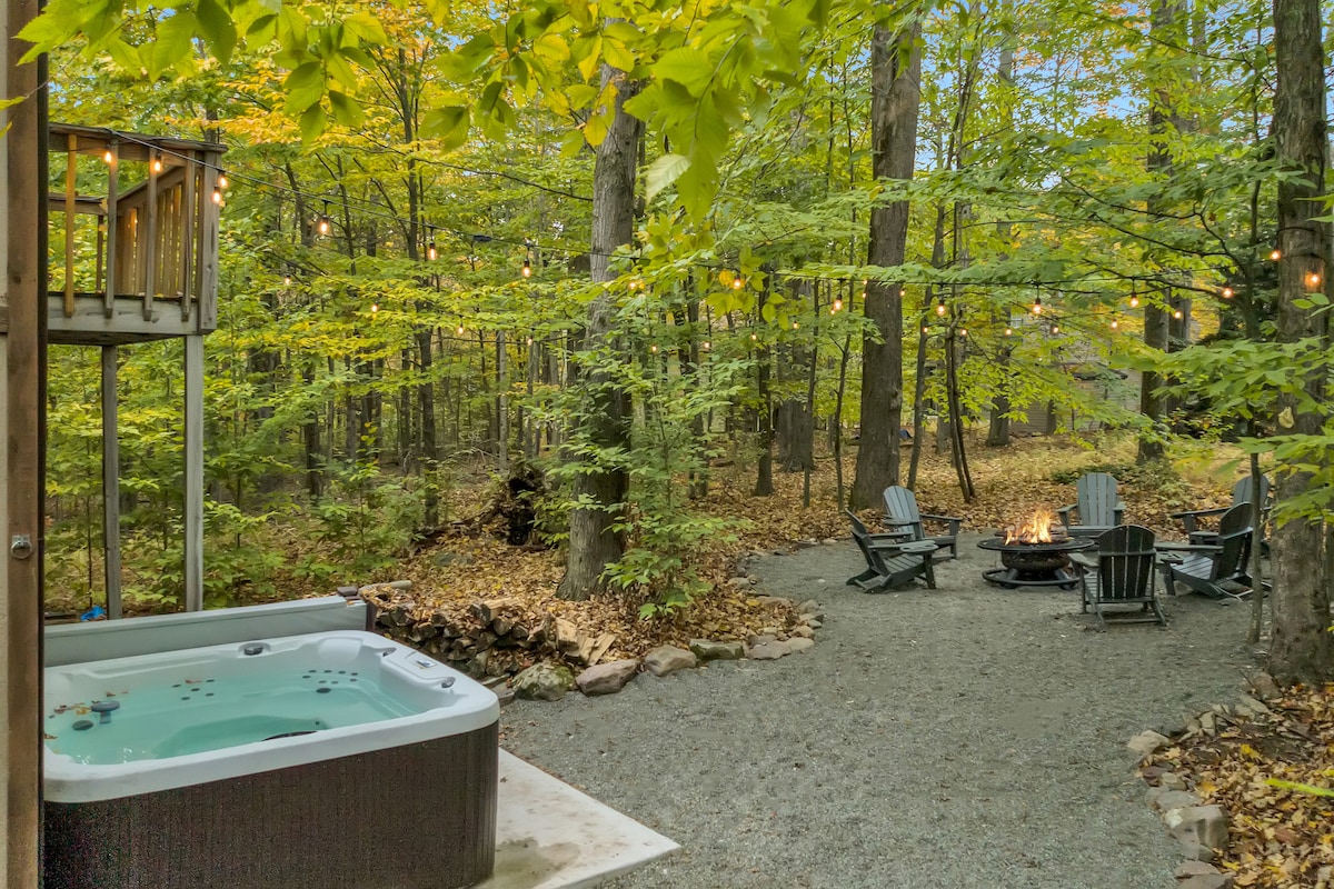 A hot tub is positioned near a circular fire pit surrounded by several Adirondack chairs, all nestled in a wooded area with vibrant green trees. Soft ambient lighting from string lights adds a warm glow to the outdoor space.