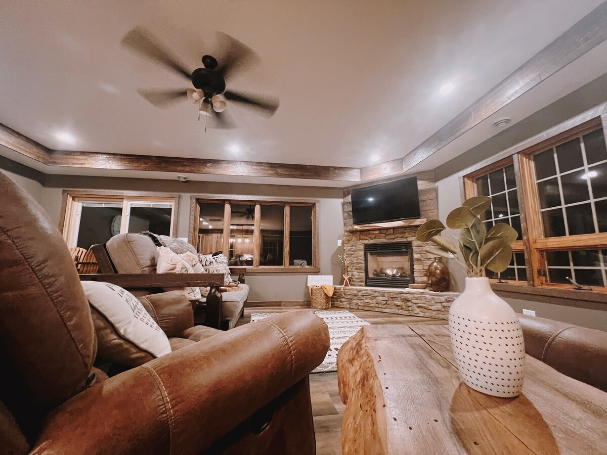A cozy living area is highlighted by a stone fireplace and a mounted television. Comfortable seating is arranged around a wooden coffee table, with large windows allowing for natural light. Neutral tones and wooden accents enhance the overall ambiance.