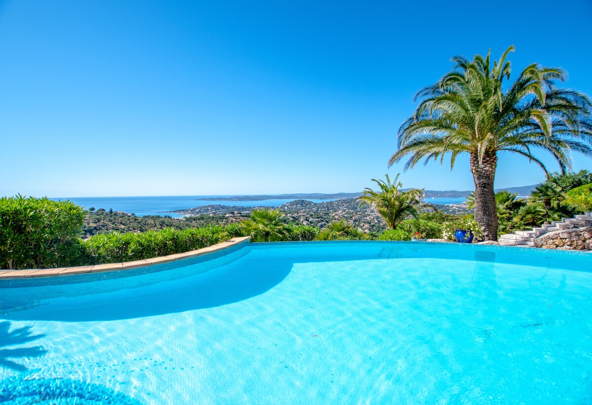 An infinity pool reflects the clear blue sky, surrounded by lush greenery and palm trees. The panoramic sea view stretches into the distance, showcasing the stunning coastline of Saint Tropez and Nartelle's beaches. The tranquil setting invites relaxation.