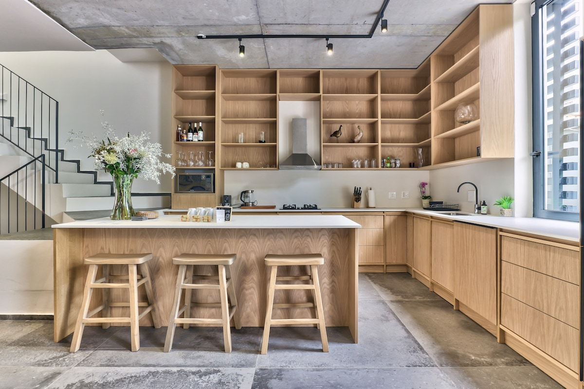 A modern kitchen features light wooden cabinetry and a central island with three stools. The sleek countertops and state-of-the-art appliances blend functionality with style. Natural light streams through the adjacent window, highlighting the open layout and clean lines of the space.