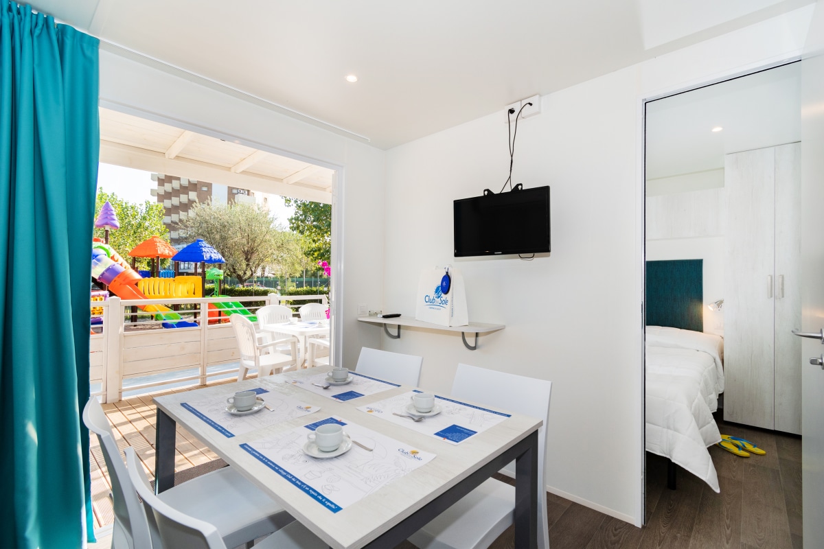 A bright dining area features a white table set for four, complemented by white chairs. A flat-screen TV is mounted on the wall. Nearby, a doorway leads to a bedroom area. Light enters through large windows adorned with blue curtains.
