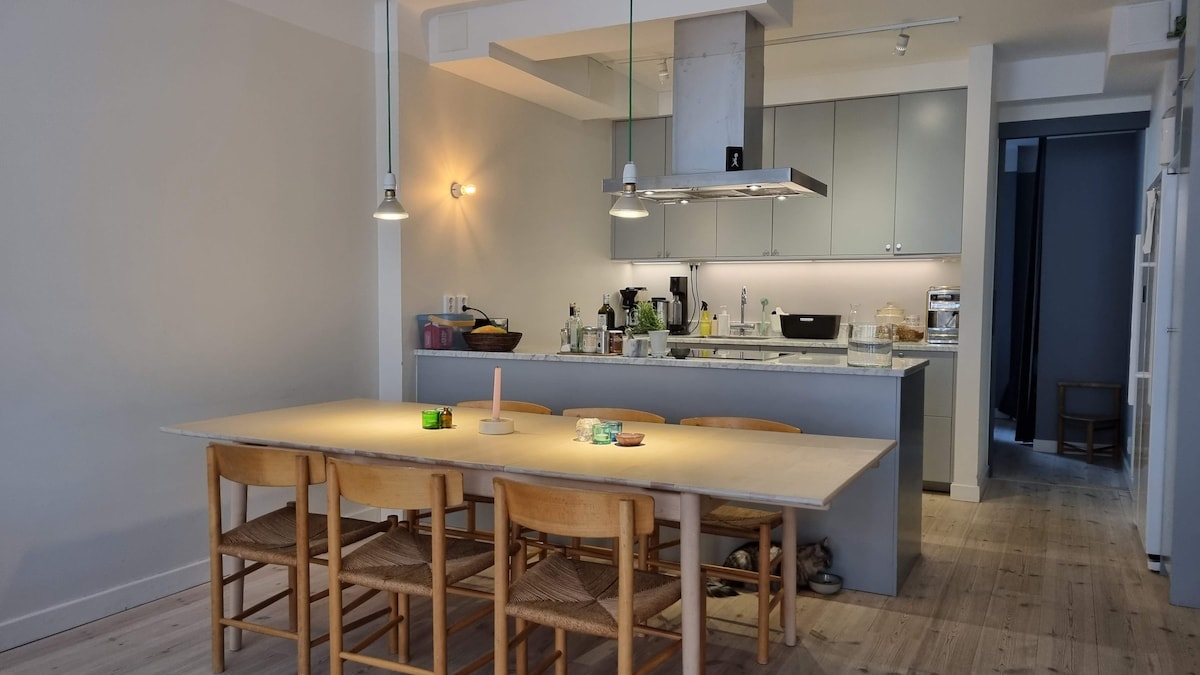 A modern kitchen is highlighted by a spacious dining table surrounded by six wooden chairs. A well-equipped countertop features various kitchen appliances, including a stove and a sink. Soft overhead lighting creates a functional space, while a doorway at the back leads to another area.