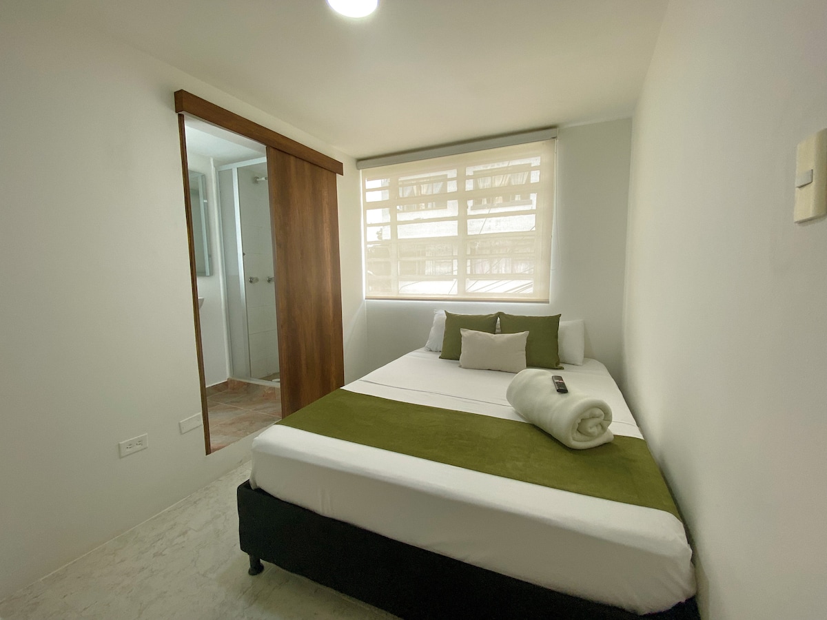 A cozy bedroom is presented with a spacious bed adorned with green and white accents. Natural light enters through a window, illuminating the clean, minimalist design. Nearby, a bathroom door is partially visible, indicating the privacy of an en-suite facility.
