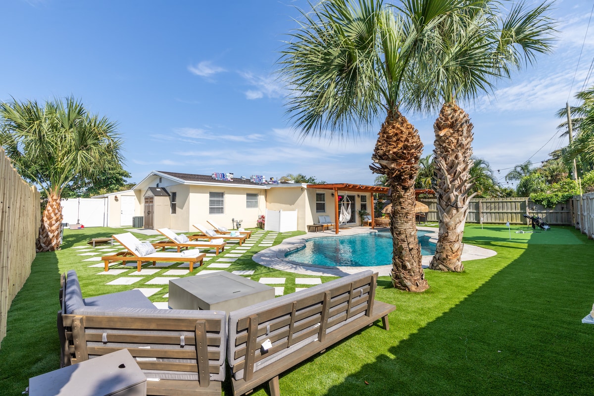 The outdoor space is characterized by a well-manicured lawn and palm trees surrounding a private saltwater pool. Lounge chairs are arranged near the water, with a shaded seating area visible under a wooden structure. A tranquil atmosphere is created with the spacious layout and vibrant greenery.