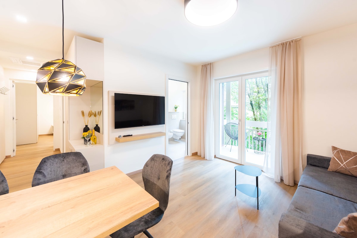 A bright living area features a comfortable sofa and a small coffee table. A dining table with four chairs sits nearby. Large windows allow natural light to fill the space, revealing a view of greenery outside. A television is mounted on the wall opposite the seating area.