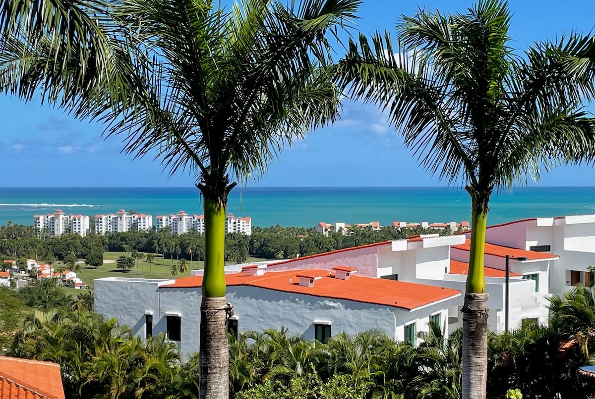 Tall palm trees frame a panoramic view of the ocean and nearby resorts. The vibrant blue water extends to the horizon, while lush greenery and red-roofed buildings fill the scenic landscape. The clear sky enhances the tranquil atmosphere of the location.