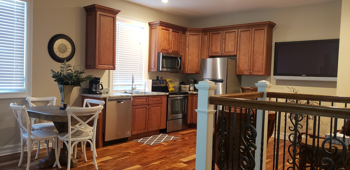 A bright kitchen area features wood cabinetry and stainless steel appliances, including a microwave and dishwasher. A circular dining table with white chairs is positioned nearby. A television mount is visible above the counter, and windows provide natural light.