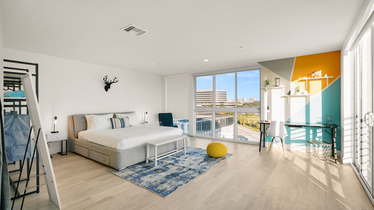 A spacious and bright bedroom is featured, showcasing a large bed with fresh bedding. Floor-to-ceiling windows allow ample natural light, providing views of the city skyline. A modern workspace with a desk is present, and vibrant wall accents add a pop of color to the neutral decor.