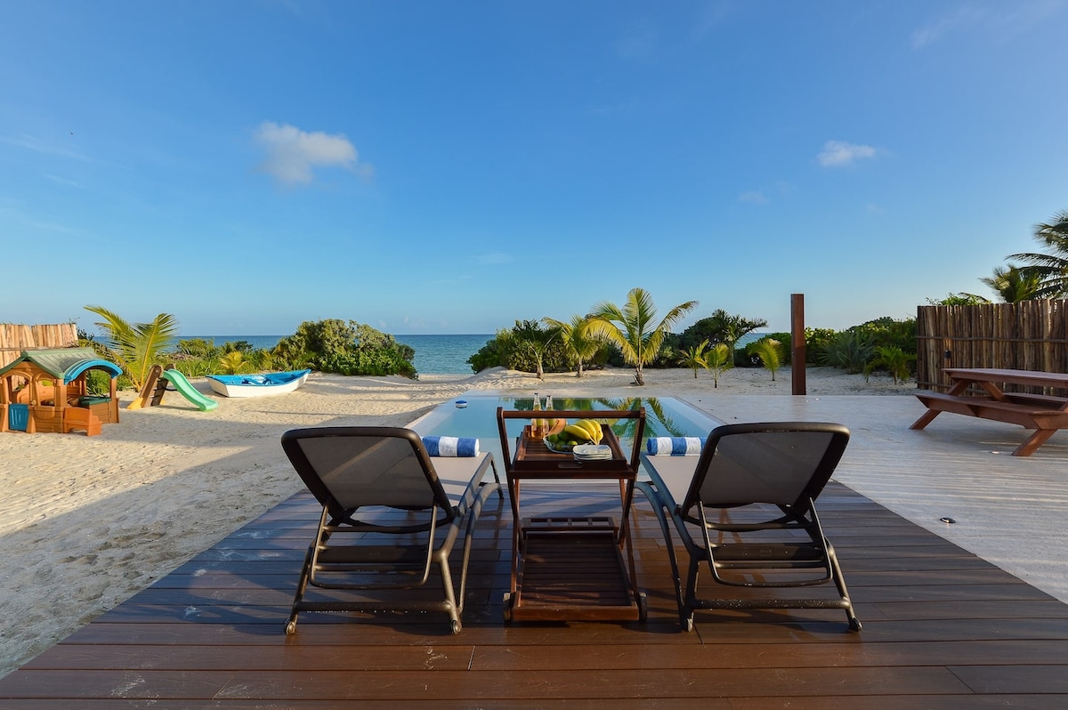 A terrace features two comfortable lounge chairs positioned beside a private pool, with clear views of the beachfront. Lush greenery borders the scene, while the tranquil sea reflects the blue sky. Tropical plants enhance the serene atmosphere, inviting relaxation and enjoyment of the coastal setting.