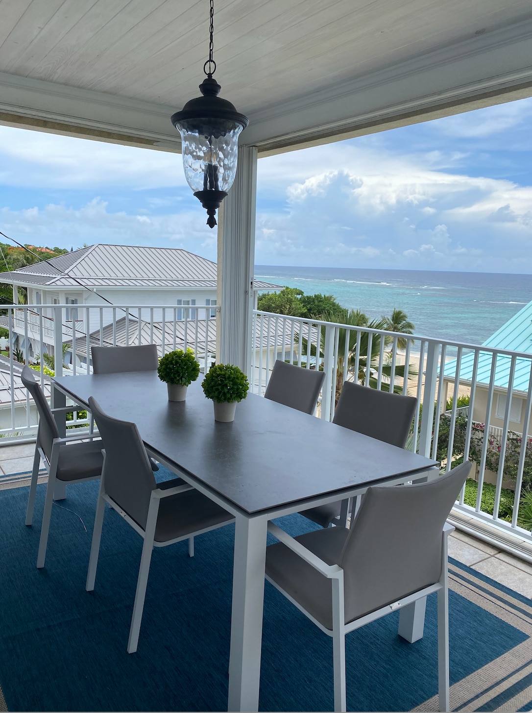 An outdoor dining area is featured on the balcony, consisting of a large table surrounded by six light-colored chairs. Two potted plants add greenery to the setting. A view of the ocean and coastline is visible in the distance, with bright skies above.