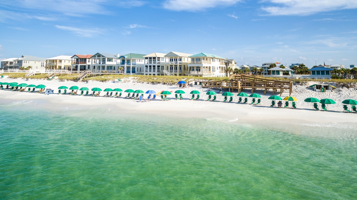 The image shows a stunning beachfront view featuring a clear, turquoise sea and soft white sand. Colorful beach umbrellas and chairs line the shore, while elegant beachfront properties are positioned in the background, emphasizing the relaxed coastal atmosphere.
