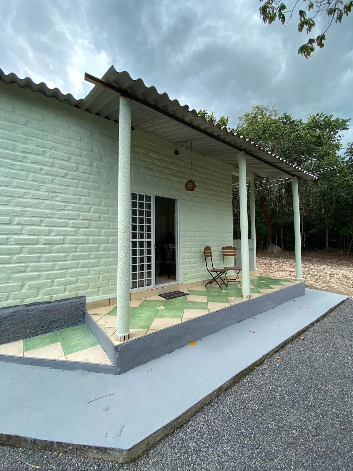 The entrance of a rustic chalet is depicted, featuring a small porch with two wooden chairs positioned on a diagonal patterned floor. The walls are painted in a soft green, and a welcoming door with glass panels leads into the interior. Surrounding greenery is visible.