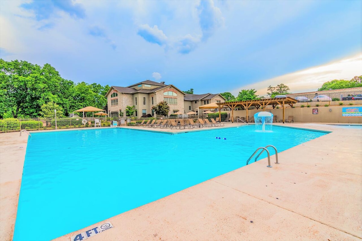 A large outdoor swimming pool is shown, featuring a bright blue water surface and a gentle waterfall feature. Surrounding the pool are lounge chairs arranged for relaxation, with shaded areas created by pergolas. Lush greenery is visible in the background, enhancing the serene atmosphere.