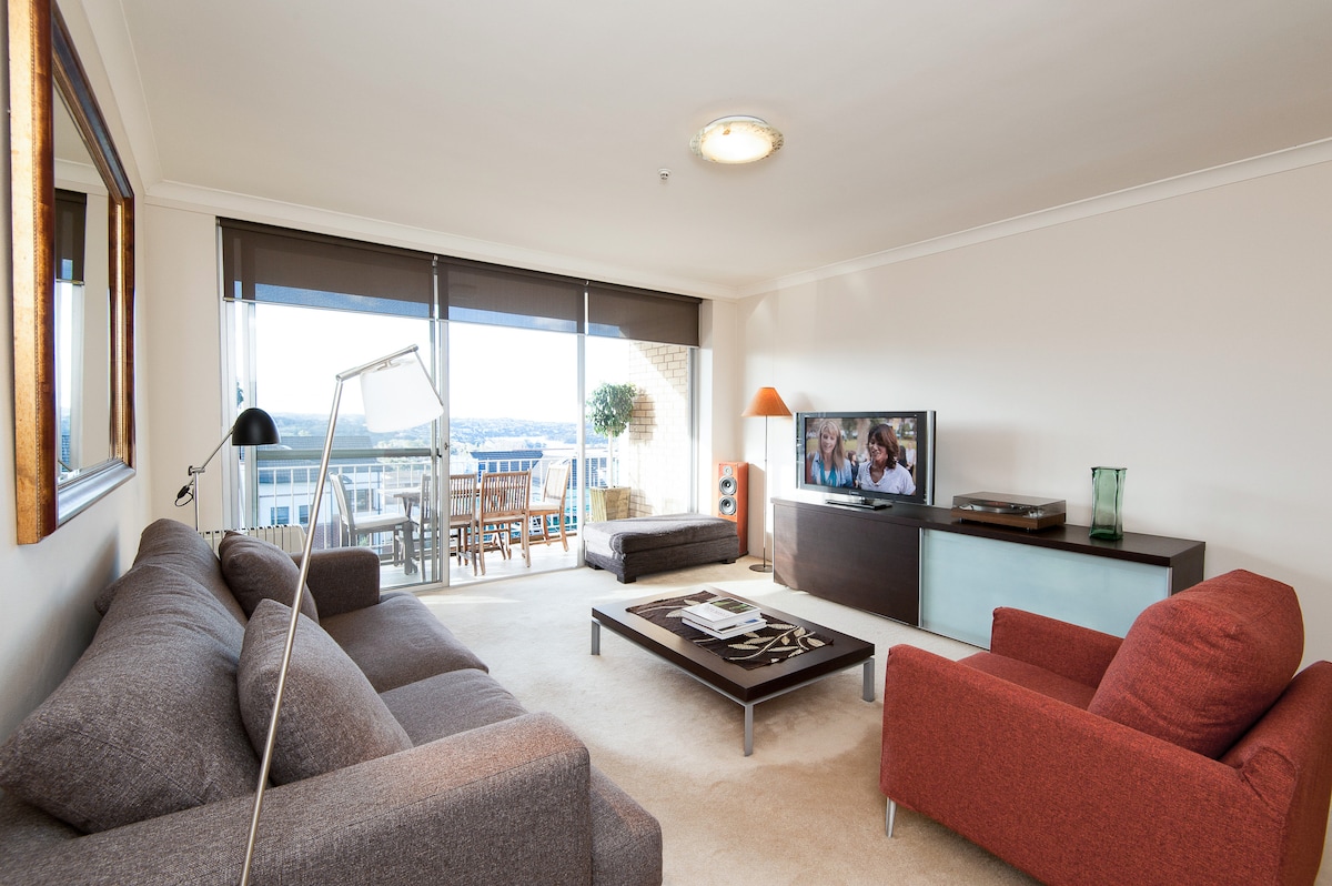 A comfortable living area is presented with a grey sectional sofa and a vibrant red armchair. A modern coffee table sits in the center, accompanied by a TV on an entertainment unit. Large glass doors lead to a balcony with outdoor seating and a view.