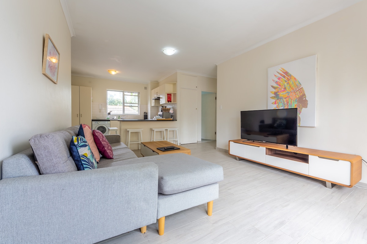 A spacious living area features a comfortable gray sofa with colorful pillows, positioned across from a modern television stand. The room is illuminated by natural light from a nearby window, and an open kitchen area is visible in the background.