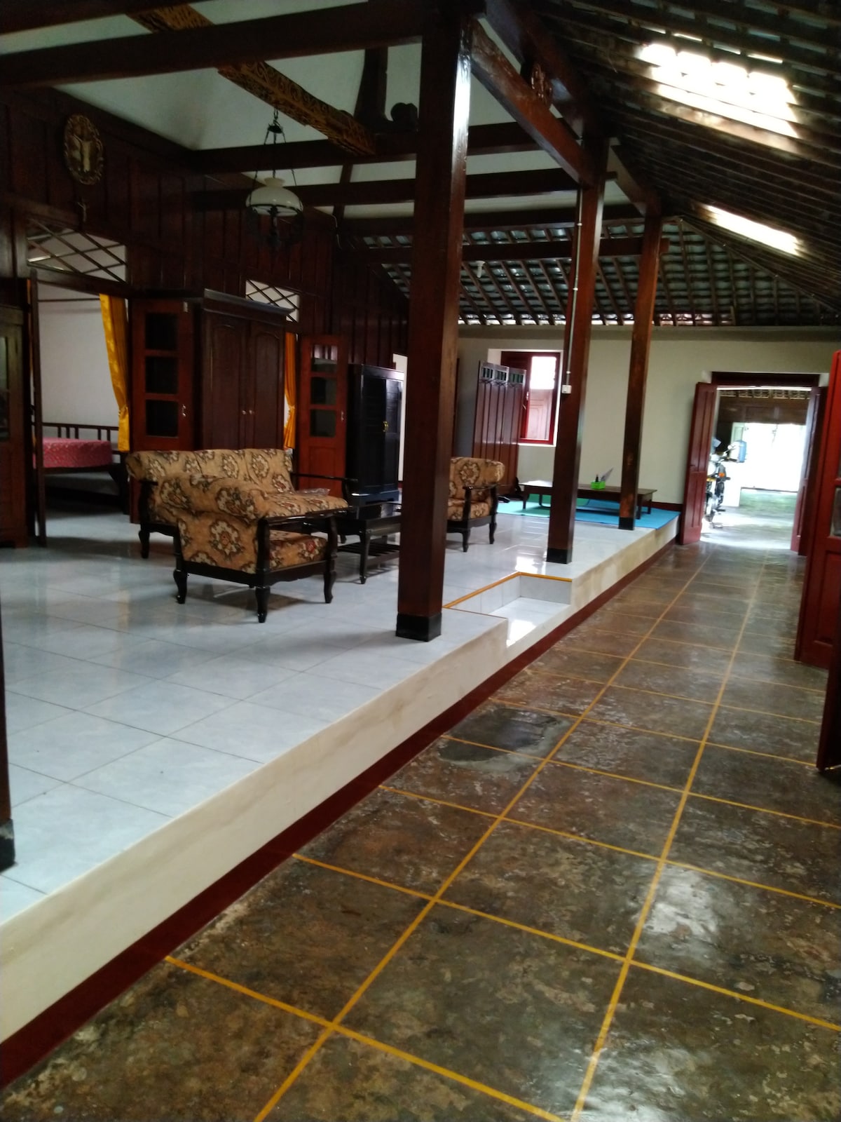 A hallway features a mix of tile and stone flooring, framed by wooden columns and walls. Two patterned armchairs are placed next to a coffee table. Doorways lead to other rooms, while natural light fills the space, enhancing the overall ambiance.