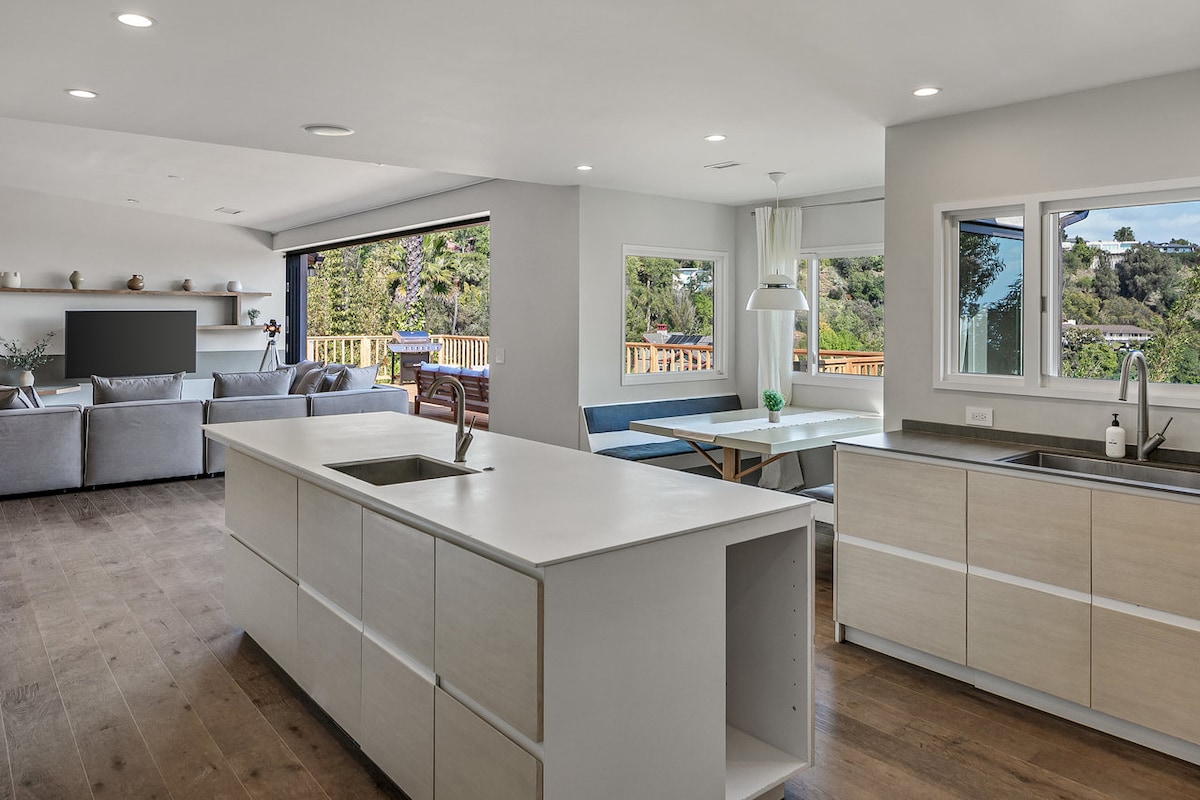 A modern kitchen is presented with a spacious center island and sleek cabinetry. Natural light floods the area through expansive windows, enhancing the open layout that connects to a comfortable living space. A smart TV is visible in the adjacent room, creating an inviting atmosphere for gatherings.