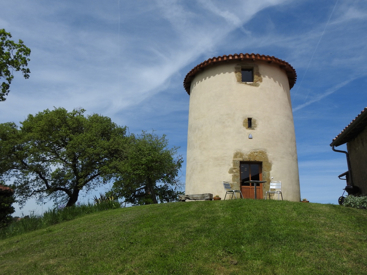 A renovated mill stands on a gently sloping grassy hill, framed by trees. The round structure features a simple wooden door and small windows, while two chairs are positioned outside, inviting relaxation. The sky above is clear with soft clouds.