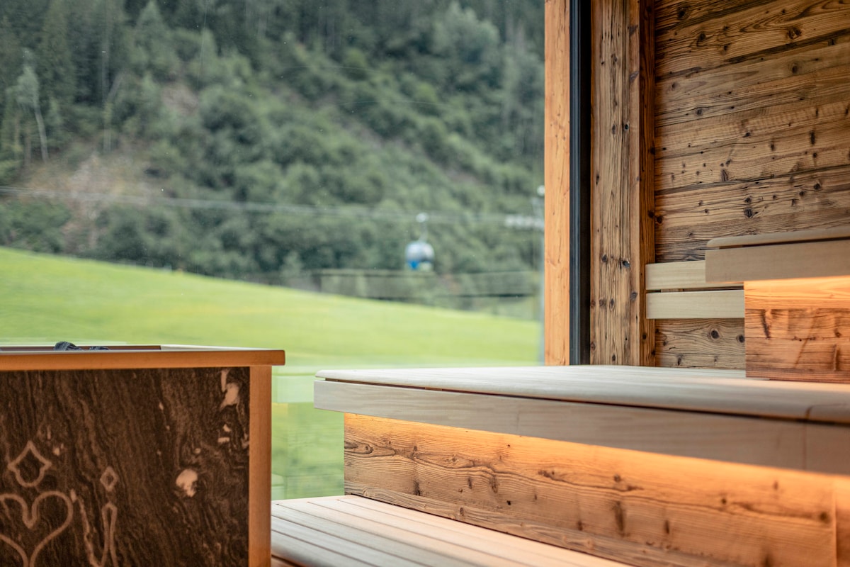 A serene sauna space is framed by wooden benches, gently illuminated. Views of green hills and a distant ski lift outside the large window create a tranquil atmosphere. Natural materials enhance the calming environment, inviting relaxation in connection with nature.