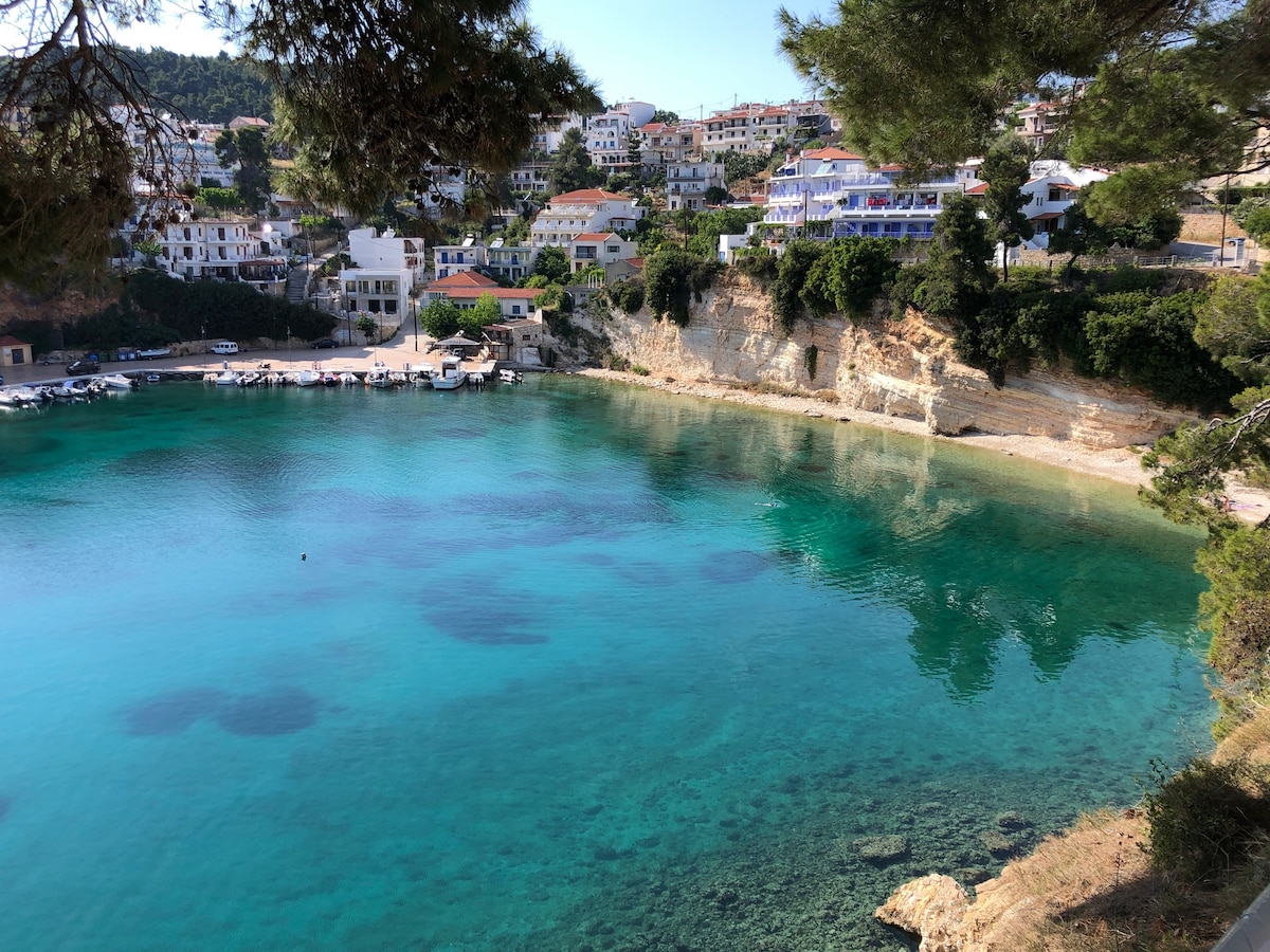 A serene cove is surrounded by rocky cliffs and lush greenery, offering a glimpse of clear turquoise waters. Several boats are anchored, and the coastal village is visible in the background, showcasing a mix of buildings nestled against the hillside.