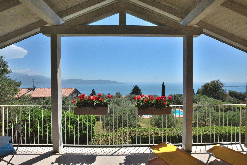 A terrace showcases a breathtaking view of the lake, framed by a railing and adorned with flowering planters. Sunlight brightens the outdoor space, which includes lounge chairs positioned for relaxation. The surrounding landscape features lush greenery and distant hills under a clear sky.